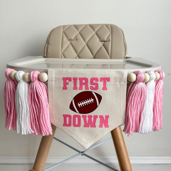 A 5 by 7 inch canvas banner with the words FIRST DOWN in pink with an image of a football between the words. There is a 5 inch pink, white and pink yarn tassel on each side with a wooden bead between each of them for a total of 6 tassels.