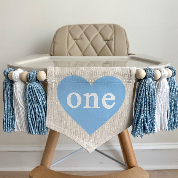 A 5 by 7 inch canvas flag with a baby blue heart and the word ONE inside the heart in white. There is a 5 inch light blue, white and light blue yarn tassel on each side, each separated by a wooden bead for a total of 6. It is hung on a high chair.