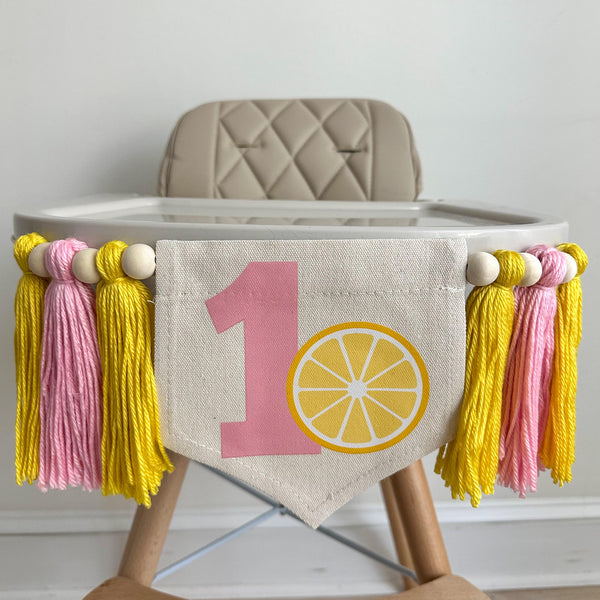 A 5 by 7 inch canvas flag with a large pink number 1 with a lemon slice beside it. There is a 5 inch yellow, pink and yellow yarn tassel on each side each separated by a wooden bead for a total of 6 tassels. It is hung on a high chair in the photo.