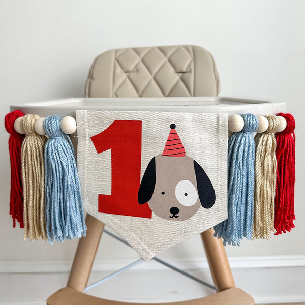 A 5 by 7 inch canvas flag with a light brown dog with a red party hat and a big red number 1 beside him. On each side there is a 5 inch light blue, tan and red yarn tassel, each separated by a wooden bead for a total of 6 tassels.
