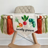 A 5 by 7 inch canvas banner with carrots, a radish and broccoli over the words LOCALLY GROWN in black. On each side there is a 5 inch dark orange, tan and sage green yarn tassel each separated by a wooden bead. It is hung on a high chair in the photo