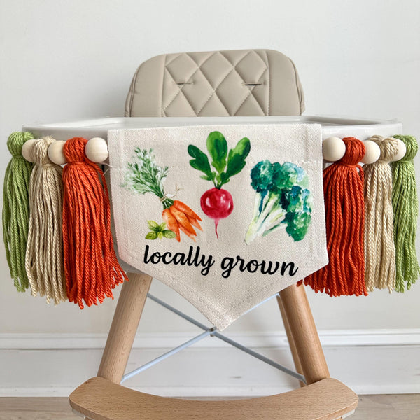A 5 by 7 inch canvas banner with carrots, a radish and broccoli over the words LOCALLY GROWN in black. On each side there is a 5 inch dark orange, tan and sage green yarn tassel each separated by a wooden bead. It is hung on a high chair in the photo