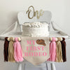 A 5 by 7 inch canvas center flag with the words MY FIRST RODEO in a pink western font and a cowboy hat in the top right corner. There are 5 inch pink, tan and brown tassels on each side each separated by a wooden bead for a total of 6 tassels.