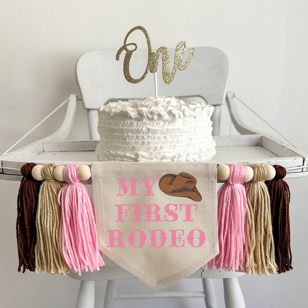 A 5 by 7 inch canvas center flag with the words MY FIRST RODEO in a pink western font and a cowboy hat in the top right corner. There are 5 inch pink, tan and brown tassels on each side each separated by a wooden bead for a total of 6 tassels.