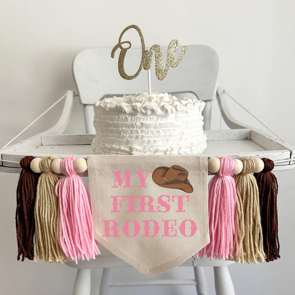 A 5 by 7 inch canvas center flag with the words MY FIRST RODEO in a pink western font and a cowboy hat in the top right corner. There are 5 inch pink, tan and brown tassels on each side each separated by a wooden bead for a total of 6 tassels.