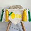 A 5 by 7 inch canvas center flag with a yellow lemon with green leaves as the O, followed by the letters NE in yellow. There is a 5 inch yellow, white and green yarn tassel on each side, each separated by a wooden bead for a total of 6 tassels.