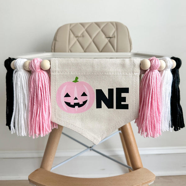 A 5 by 7 inch canvas flag with a pink jack o lantern followed by the letters NE in black. There is a pink, white and black 5 inch yarn tassel on each side, separated by wooden beads. It is hung on a high chair in the photo.