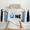 A 5 x 7 inch canvas center flag with a light blue Jack O Lantern followed by the letters NE in black. On each side there is a light blue, white and black 5 inch yarn tassel, each separated by a wooden bead. It is hung on a high chair in the photo.