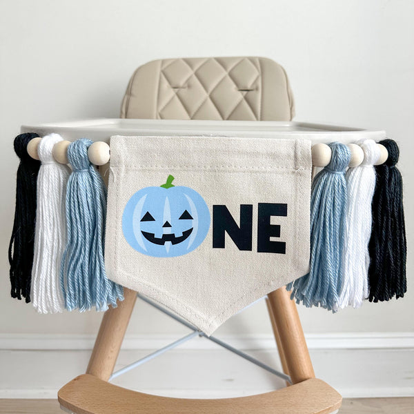 A 5 x 7 inch canvas center flag with a light blue Jack O Lantern followed by the letters NE in black. On each side there is a light blue, white and black 5 inch yarn tassel, each separated by a wooden bead. It is hung on a high chair in the photo.
