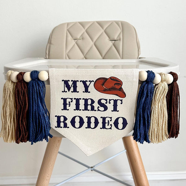 A 5 by 7 inch canvas banner with the words MY FIRST RODEO in a navy western font with a cowboy hat accent. There is a 5 inch navy blue, tan and brown yarn tassel on each side with a wooden bead between each of them for a total of 6 tassels.