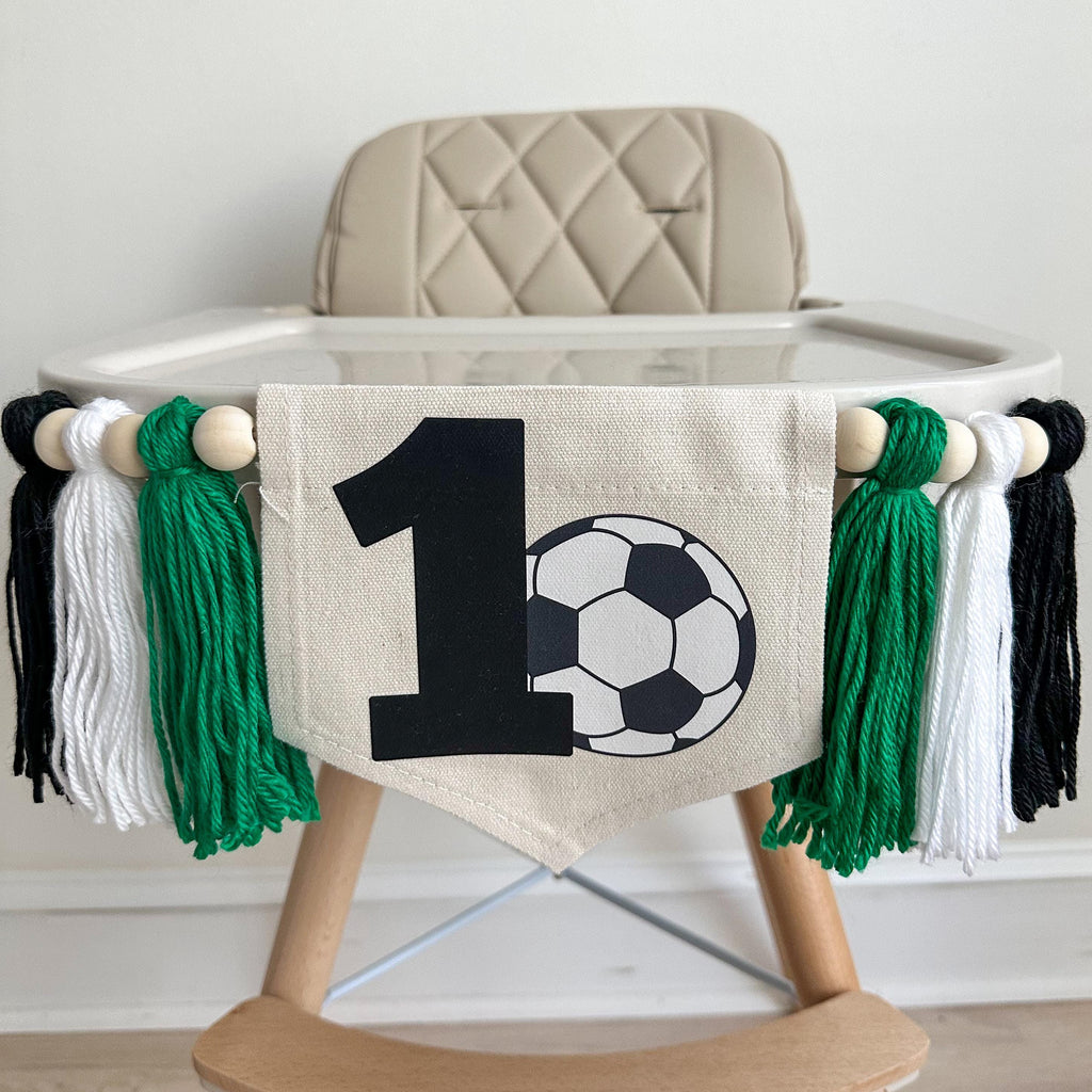 A 5 by 7 inch canvas center flag with a black number 1 and a soccer ball beside it. On each side there is a 5 inch kelly green, white and black yarn tassel, each separated by a wooden bead for a total of 6 tassels. It is displayed on a high chair.