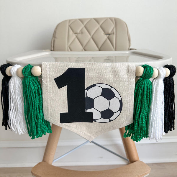 A 5 by 7 inch canvas center flag with a black number 1 and a soccer ball beside it. On each side there is a 5 inch kelly green, white and black yarn tassel, each separated by a wooden bead for a total of 6 tassels. It is displayed on a high chair.