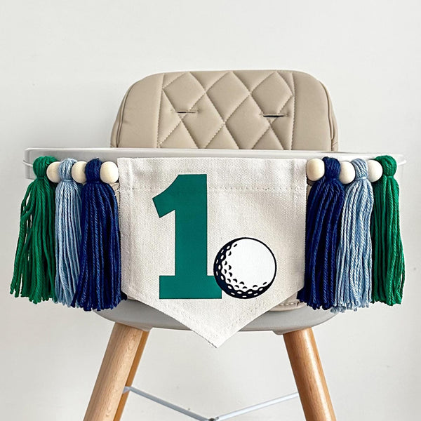A 5 by 7 inch canvas center flag with a large green number 1 with a golf ball beside it. There is a navy, light blue and kelly green 5 inch yarn tassel on each side, each separated by a wooden bead for a total of 6 tassels and 6 beads.