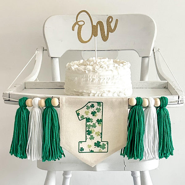 A 6 x 7 inch canvas center flag with a number 1 outlined in green full of clovers in different colors. There is a green, white and green 5 inch yarn tassel on each side each separated by a wooden bead. It is hung on a high chair in the photo.