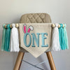 A 6 by 7 inch canvas center flag with the word ONE in light aqua and bunny ears on the O. On each side there is a 5 inch white, light aqua and soft green yarn tassel each separated by a wooden bead for a total of 6 tassels.