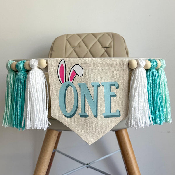 A 6 by 7 inch canvas center flag with the word ONE in light aqua and bunny ears on the O. On each side there is a 5 inch white, light aqua and soft green yarn tassel each separated by a wooden bead for a total of 6 tassels.