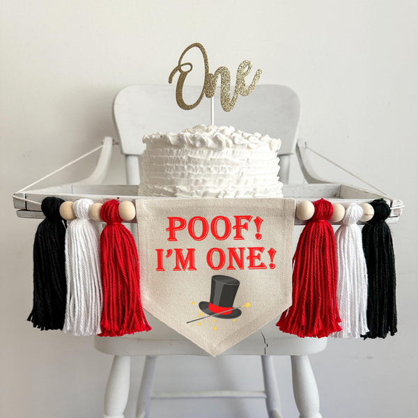 A 6 by 7 inch canvas center flag with words POOF I&#39;M ONE in red and a magician&#39;s hat under the words. On each side there is a 5 inch red, white and black yarn tassel each separated by a wooden bead for a total of 6 tassels.