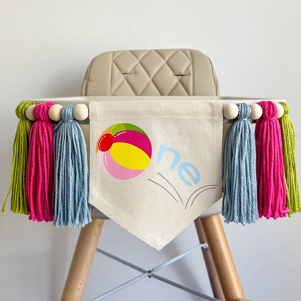 A 6 by 7 inch canvas center flag with a colorful bouncing beach ball and the letters NE in light blue. On each side there is a 5 inch light blue, hot pink and lime green yarn tassel each separated by a wooden bead for a total of 6 tassels.