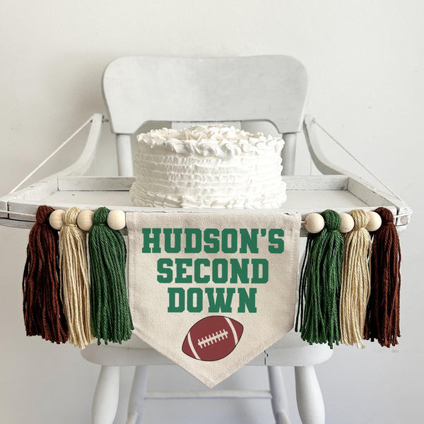 A 6 by 7 inch canvas center flag with the words CUSTOM&#39;S SECOND DOWN in green and a football under the words. On each side there is a 5 inch dark green, tan and brown yarn tassel each separated by a wooden bead for a total of 6 tassels.