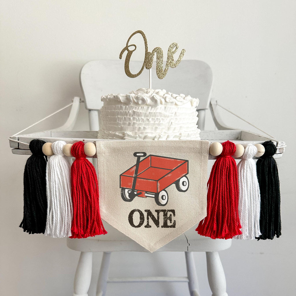A 5 by 7 inch canvas center flag with a little red wagon and the word ONE in black. There are 5 inch red, white and black yarn tassels on each side for a total of 6 tassels. The banner is shown on a high chair.