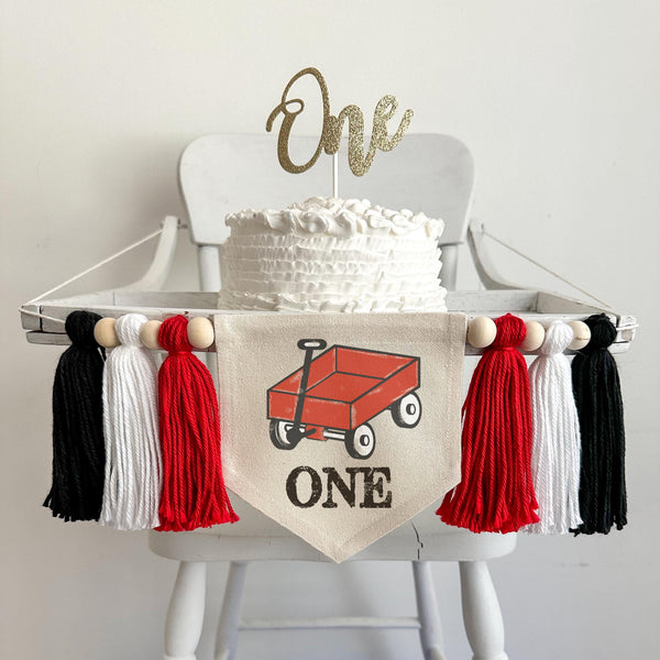 A 5 by 7 inch canvas center flag with a little red wagon and the word ONE in black. There are 5 inch red, white and black yarn tassels on each side for a total of 6 tassels. The banner is shown on a high chair.