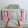 A 5 by 7 inch canvas center flag with a white snowflake surrounded by pink and the letters NE in white also surrounded by pink. There is a 5 inch pink, white and light gray yarn tassel on each side, each separated by a wooden bead for a total of 6.