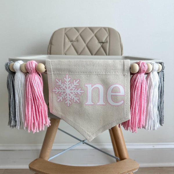 A 5 by 7 inch canvas center flag with a white snowflake surrounded by pink and the letters NE in white also surrounded by pink. There is a 5 inch pink, white and light gray yarn tassel on each side, each separated by a wooden bead for a total of 6.