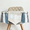 A 5 by 7 inch canvas center flag with a white snowflake surrounded by baby blue and the letters NE in white also surrounded by baby blue. There is a 5 inch baby blue, white and light gray yarn tassel on each side, each separated by a wooden bead.