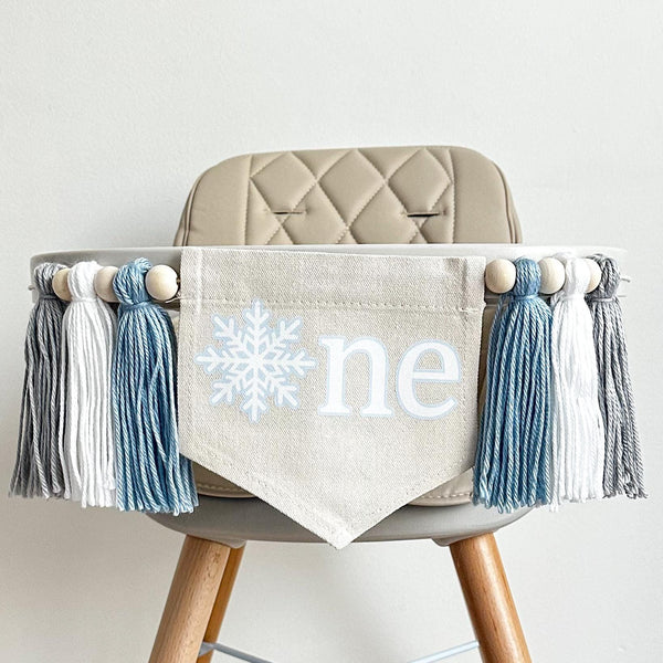 A 5 by 7 inch canvas center flag with a white snowflake surrounded by baby blue and the letters NE in white also surrounded by baby blue. There is a 5 inch baby blue, white and light gray yarn tassel on each side, each separated by a wooden bead.