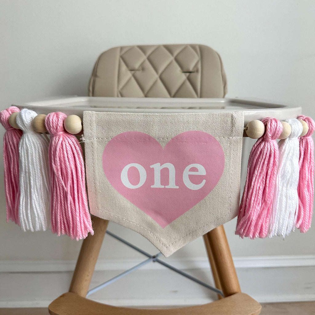A 5 by 7 inch canvas center flag with a pink heart and the word ONE inside the heart. There is a pink, white and pink 5 inch yarn tassel on each side, each separated by a wooden bead for a total of 6 tassels. It is hung on a white high chair.