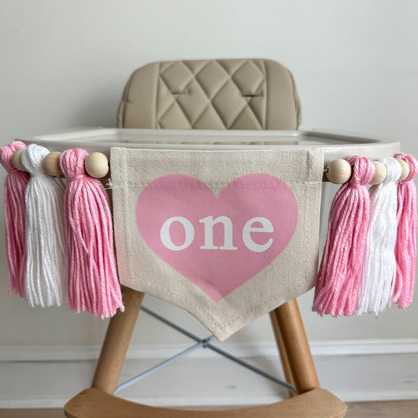 A 5 by 7 inch canvas center flag with a pink heart and the word ONE inside the heart. There is a pink, white and pink 5 inch yarn tassel on each side, each separated by a wooden bead for a total of 6 tassels. It is hung on a white high chair.
