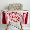 A 5 by 7 inch center flag with the word ONE in dark pink script. There are a lot of tiny hearts in red and pink around the word to form the shape of a big heart. There are 5 inch pink, red and white yarn tassels on each side separated by wooden beads