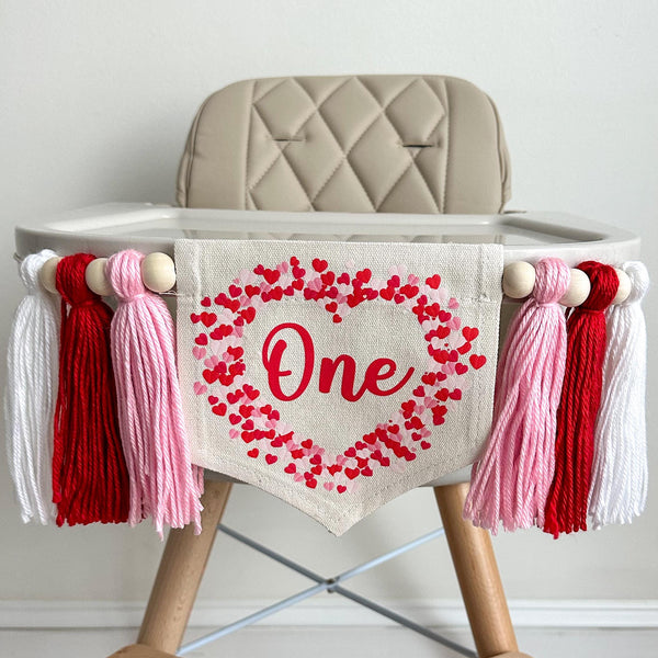 A 5 by 7 inch center flag with the word ONE in dark pink script. There are a lot of tiny hearts in red and pink around the word to form the shape of a big heart. There are 5 inch pink, red and white yarn tassels on each side separated by wooden beads