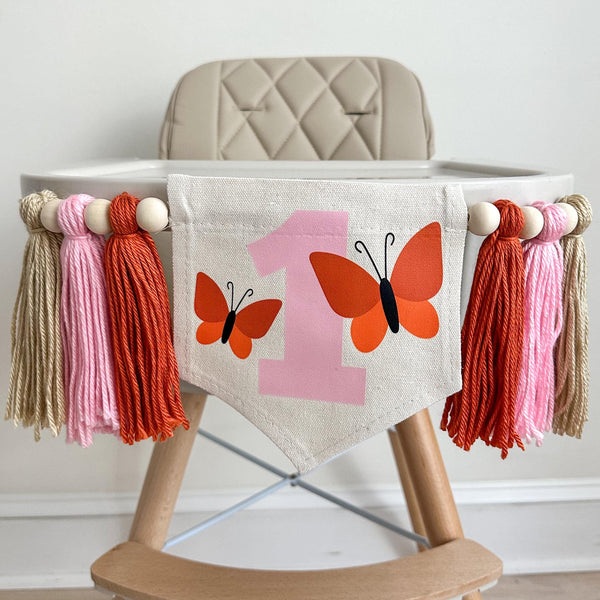 This is a 6.5 x 7 inch canvas flag with a large pink 1 in the middle. There is an orange butterfly on either side of the pink 1. On each side of the flag there are 3 tassels in dark orange, pink and white with wooden beads in between each tassel.