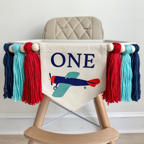 A 5 by 7 inch canvas flag with a blue, teal and red vintage plane and the word ONE in navy above the plane. There is a 5 inch red, light aqua and navy yarn tassel on each side, each separated by a wooden bead for a total of 6 tassels.