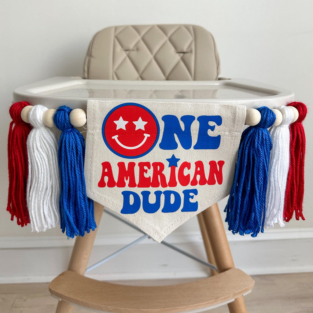 A 5 by 7 inch canvas flag with the words ONE AMERCAN DUDE in red and blue with a red smile face as the O in one. On each side there is a 5 inch royal blue, white and red yarn tassel each separated by a wooded bead for a total of 6 tassels.
