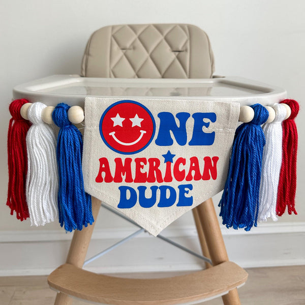 A 5 by 7 inch canvas flag with the words ONE AMERCAN DUDE in red and blue with a red smile face as the O in one. On each side there is a 5 inch royal blue, white and red yarn tassel each separated by a wooded bead for a total of 6 tassels.