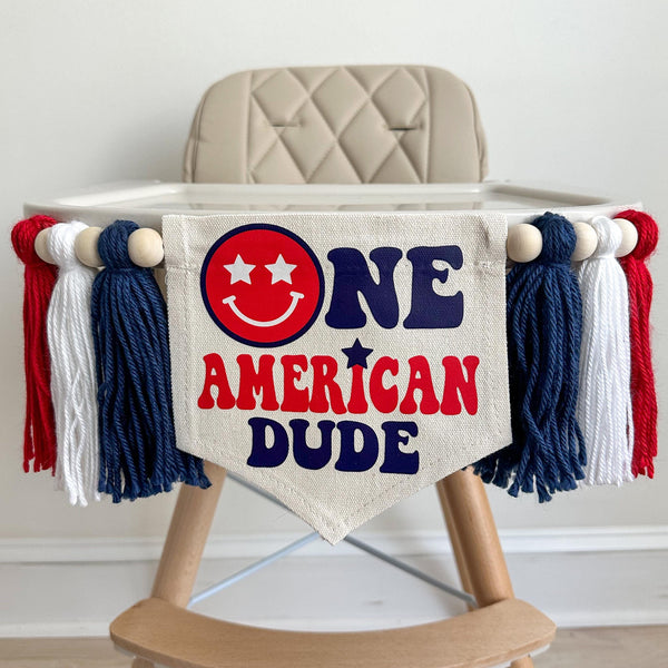 A 5 by 7 inch canvas flag with the words ONE AMERICAN DUDE in red and navy blue with a red smiling face as the O in one. On each side there is a 5 inch navy blue, white and red yarn tassel each separated by a wooden bead for a total of 6 tassels.