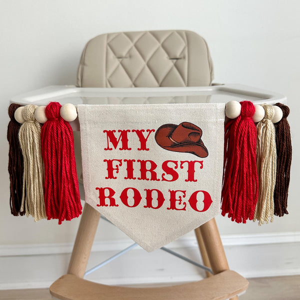 A 5 by 7 inch canvas banner with the words MY FIRST RODEO in a red western style font with a cowboy hat accent. There is a 5 inch red, tan and brown yarn tassel on each side with a wooden bead between each of them for a total of 6 tassels.