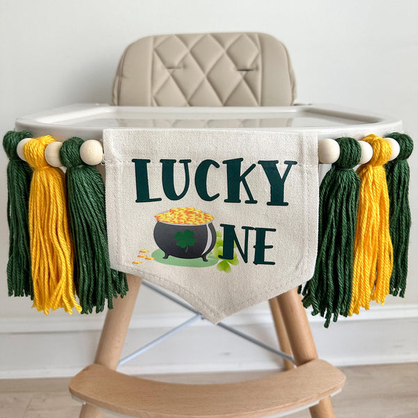 A 5 by 7 inch canvas center flag with the words LUCKY ONE in dark green with a pot of gold coins as the letter O. On each side there is a 5 inch dark green, golden yellow and dark green yarn tassel each separated by a wooden bead for a total of 6.