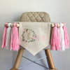A 6 by 7 inch canvas center flag with a bunny inside a floral wreath to make the letter O followed by NE in pink.  On each side there is a 5 inch pink, white and pink yarn tassel each separated by a wooden bead for a total of 6 tassels.