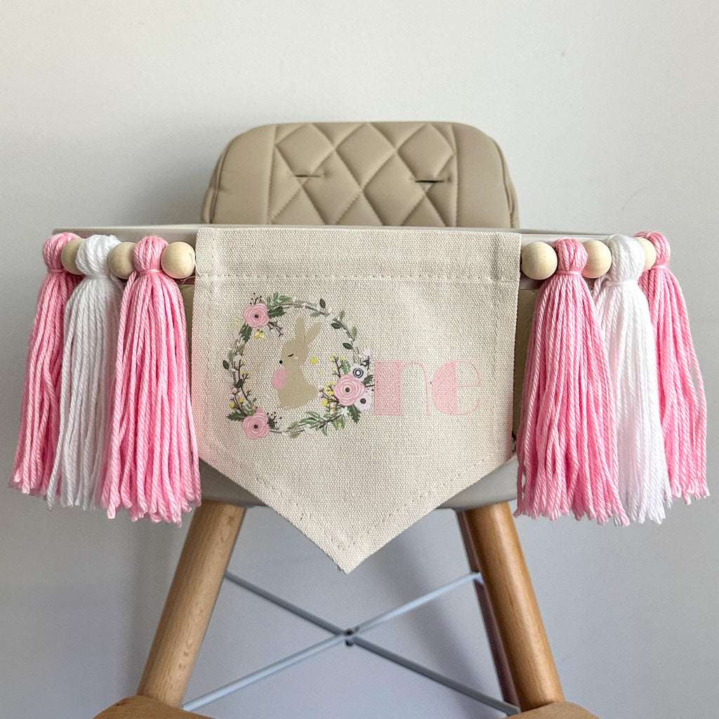 A 6 by 7 inch canvas center flag with a bunny inside a floral wreath to make the letter O followed by NE in pink.  On each side there is a 5 inch pink, white and pink yarn tassel each separated by a wooden bead for a total of 6 tassels.