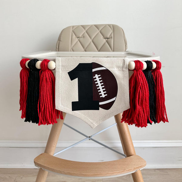 A 6 by 7 inch canvas center flag with a black number 1 and a brown football. On each side there is a 5 inch red, black and red yarn tassel each separated by a wooden bead for a total of 6 tassels. It is hung on a high chair in the photo.
