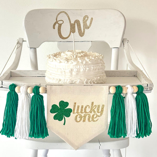 A 5 by 7 inch canvas center flag with the words LUCKY ONE in gold glitter script and a green glitter clover beside the words. On each side there is a 5 inch green, white and green yarn tassel each separated by a wooden bead for a total of 6 tassels.