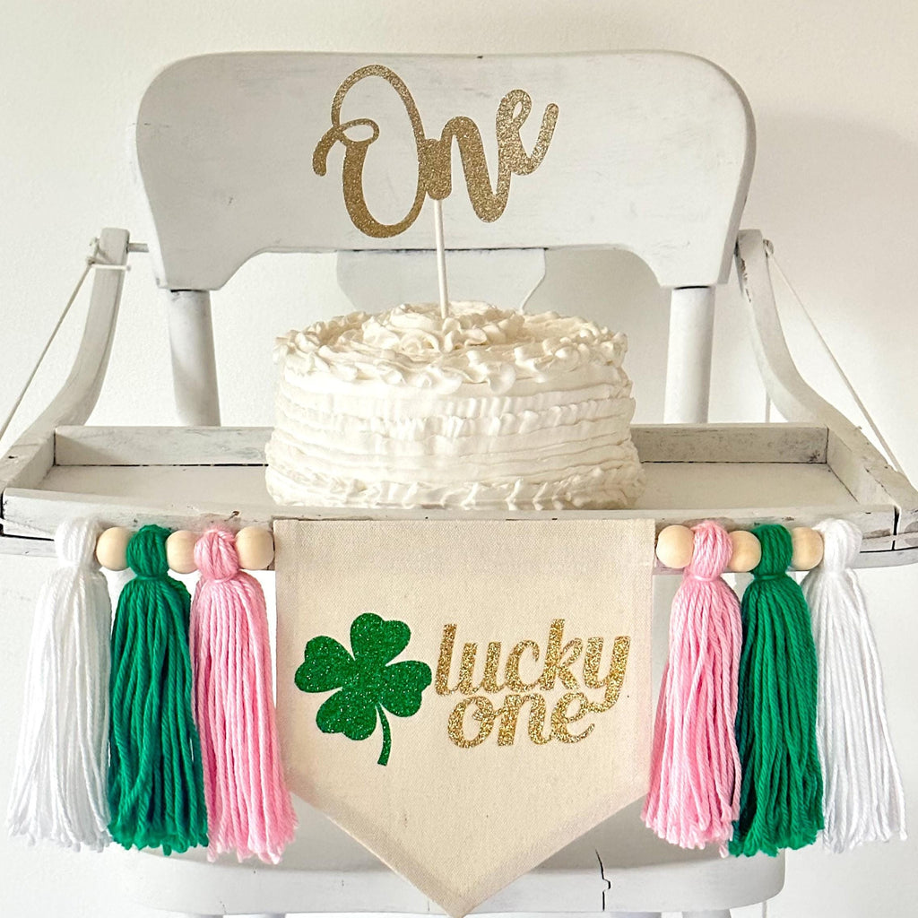 A 5 by 7 inch canvas center flag with the words LUCKY ONE in gold glitter script and a green glitter clover beside the words. On each side there is a 5 inch pink, green and white yarn tassel each separated by a wooden bead for a total of 6 tassels.