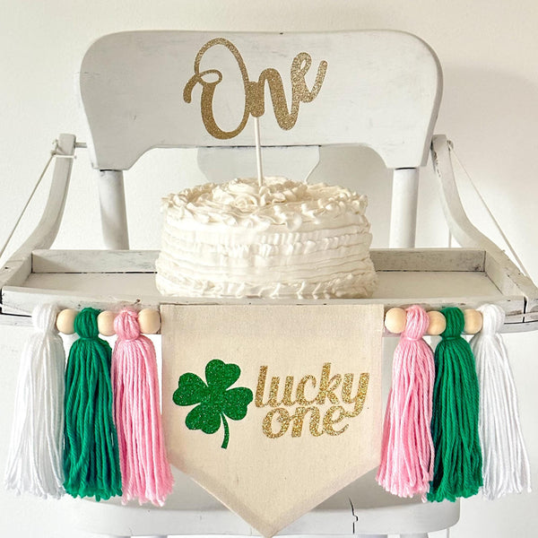 A 5 by 7 inch canvas center flag with the words LUCKY ONE in gold glitter script and a green glitter clover beside the words. On each side there is a 5 inch pink, green and white yarn tassel each separated by a wooden bead for a total of 6 tassels.