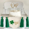 A 5 by 7 inch canvas center flag with a green glitter clover beside the gold script letters NE to spell ONE. On each side there is a 5 inch green, white and green yarn tassel each separated by a wooden bead for a total of 6 tassels.