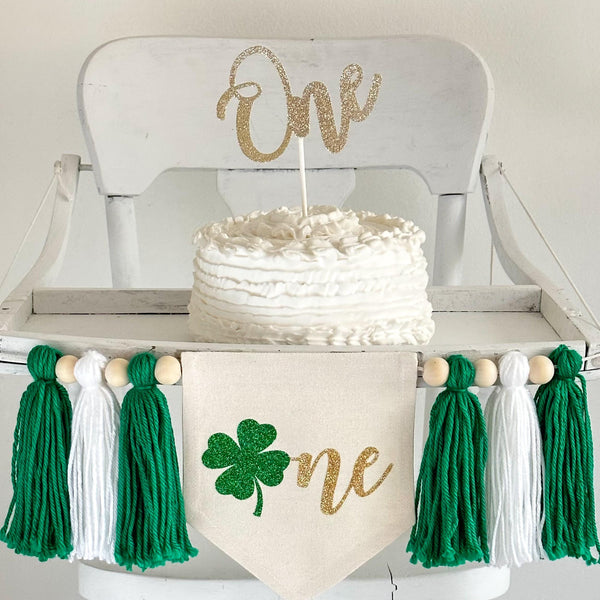A 5 by 7 inch canvas center flag with a green glitter clover beside the gold script letters NE to spell ONE. On each side there is a 5 inch green, white and green yarn tassel each separated by a wooden bead for a total of 6 tassels.