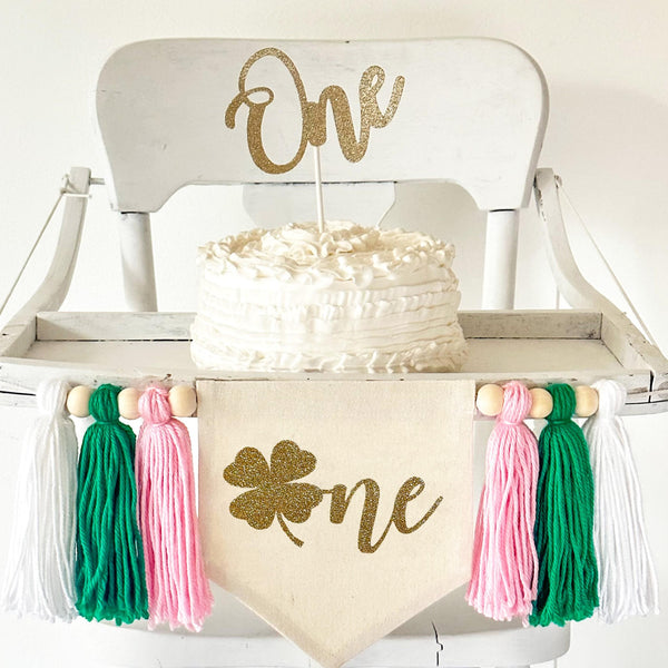 A 5 by 7 inch canvas center flag with a gold glitter clover beside the gold glitter letters NE to spell the word ONE. On each side there is a 5 inch pink, green and white yarn tassel each separated by a wooden bead for a total of 6 tassels.