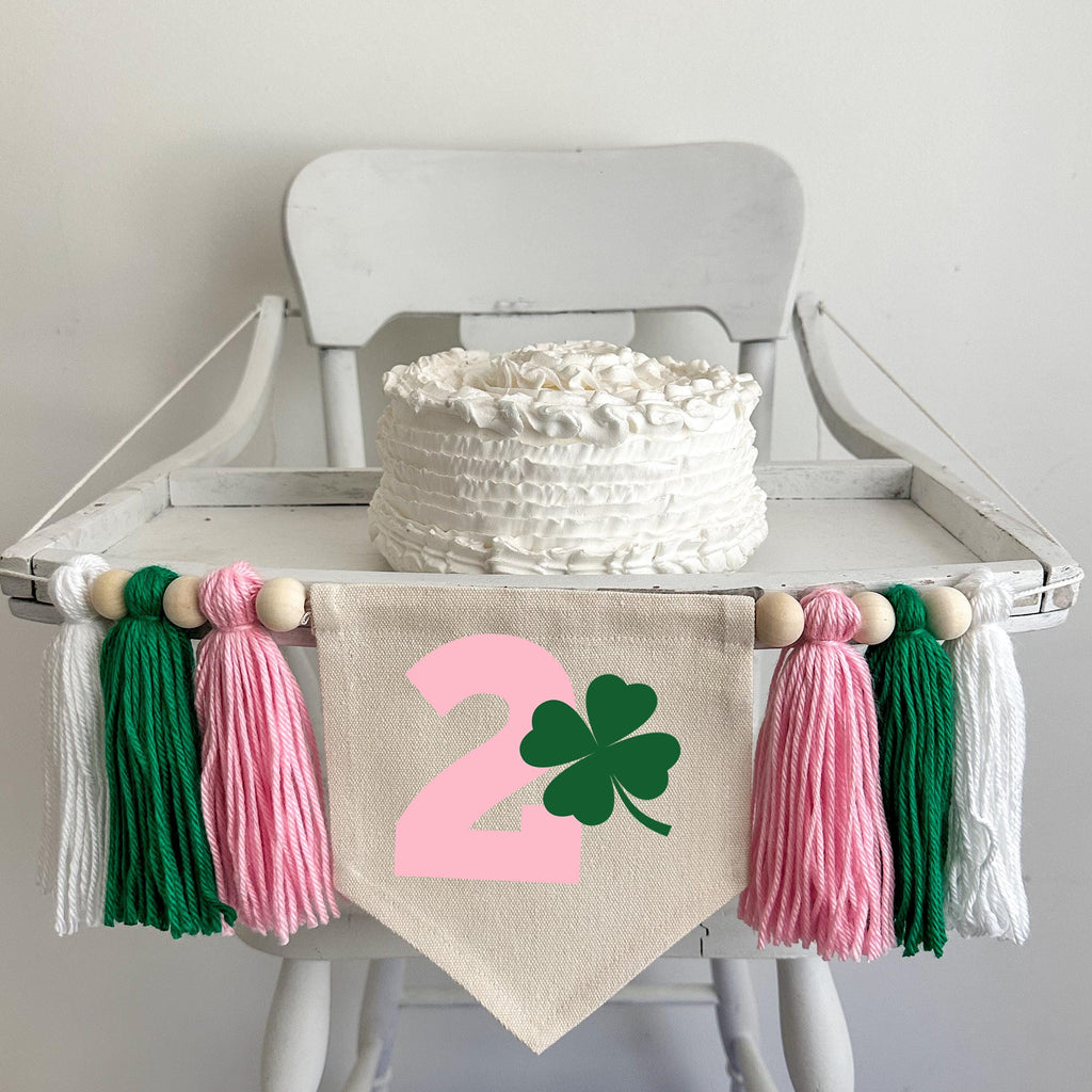 A 5 by 7 inch canvas center flag with a printed pink number 2 beside a green clover. There is a 5 inch pink, green and white yarn tassel on each side, each separated by a wooden bead for a total of 6. The banner is strung on a high chair in the photo